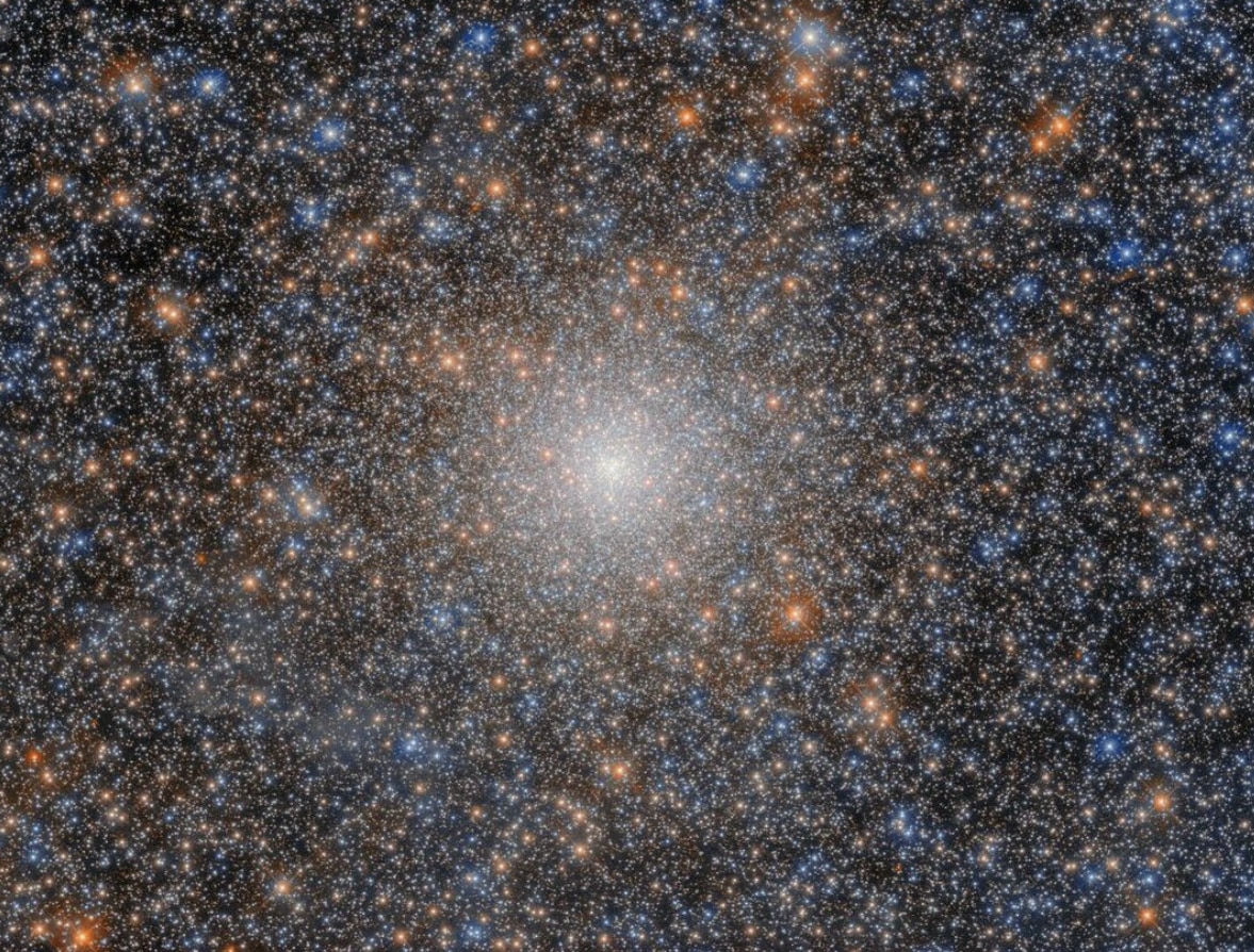 A globular cluster that looks like a very dense, ball-shaped collection of many shining stars in colors of white, yellow-orange, and blue. Some stars appear a bit larger and brighter than others, with the brightest having faint cross-shaped diffraction spikes. The cluster’s stars are scattered mostly uniformly, with their density increasing toward the cluster’s core where they merge into a strong, bright-white glow.⁣ 
