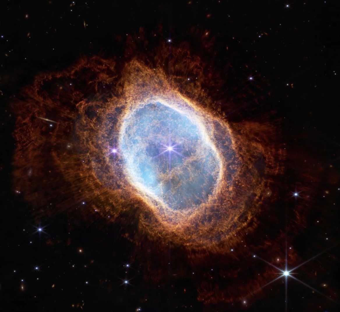 A planetary nebula, seen by the Webb telescope's NIRCam instrument, against the blackness of space, with points of starlight behind it. The nebula itself is shaped like an irregular oval, with lacy, reddish orange plumes of gas and dust.