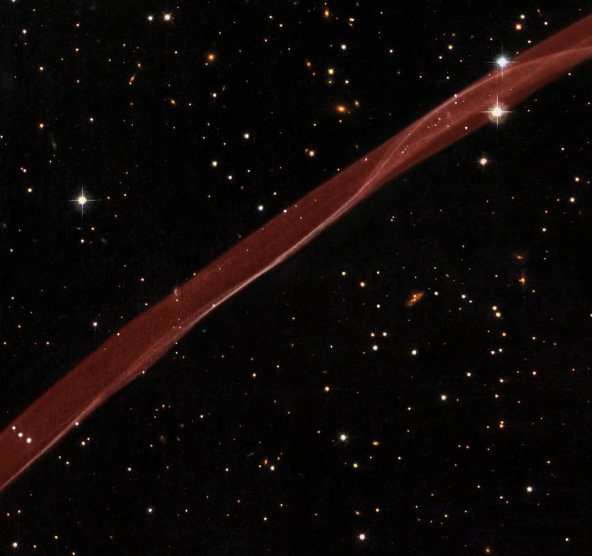 A thin, red ribbon of gas crosses diagonally over the scene. Details in the trail show dimension and twisting of the stream of matter. In the background, black space is dotted with yellow stars and galaxies.